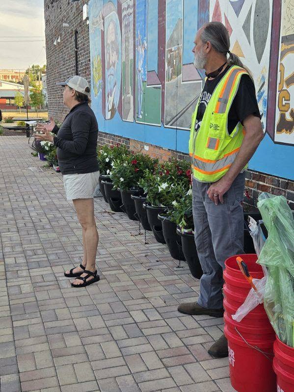 A woman and a man in a safety vest stand beside a mural wall and several potted plants on a brick walkway.