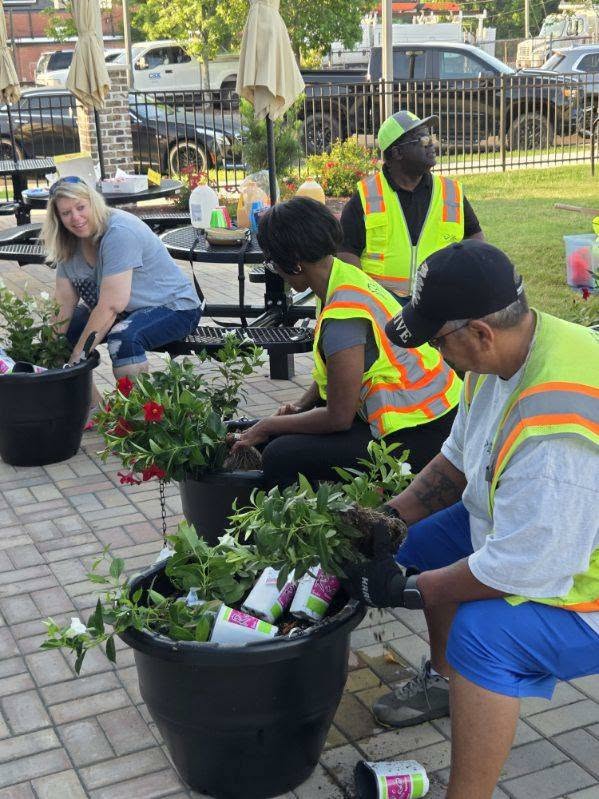 Several people wearing safety vests are planting flowers in large pots outdoors, sitting at picnic tables and on the pavement, participating in a community gardening activity.