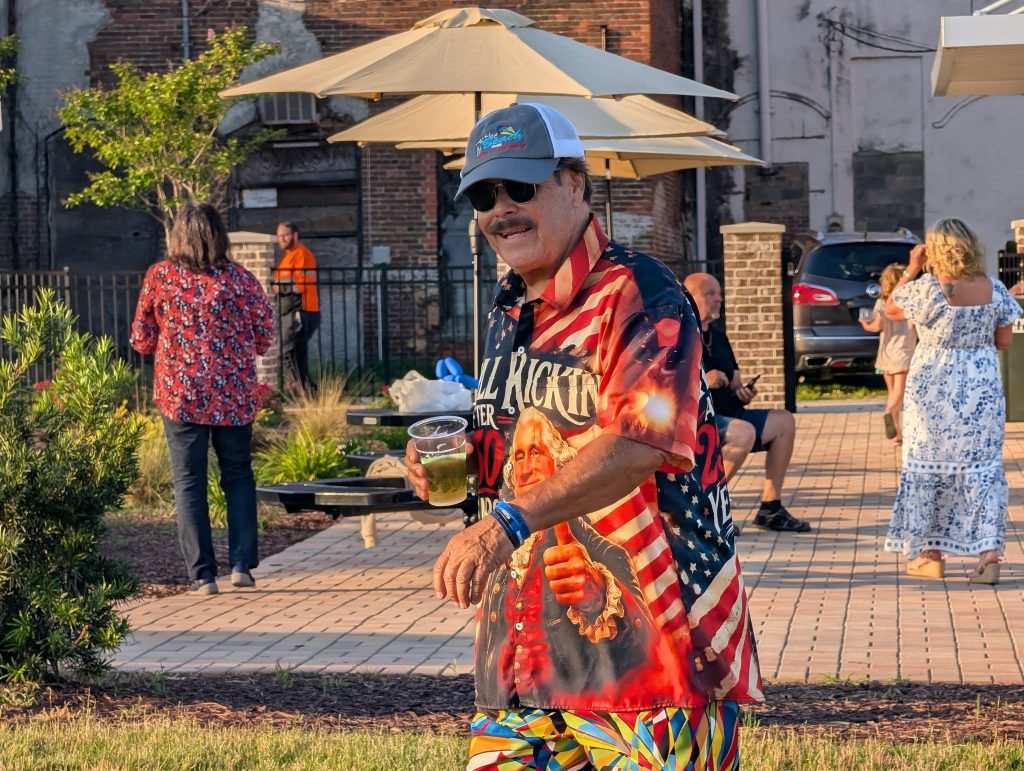 Man wearing a colorful shirt and sunglasses holding a drink outdoors at a casual event, with people and umbrellas in the background.