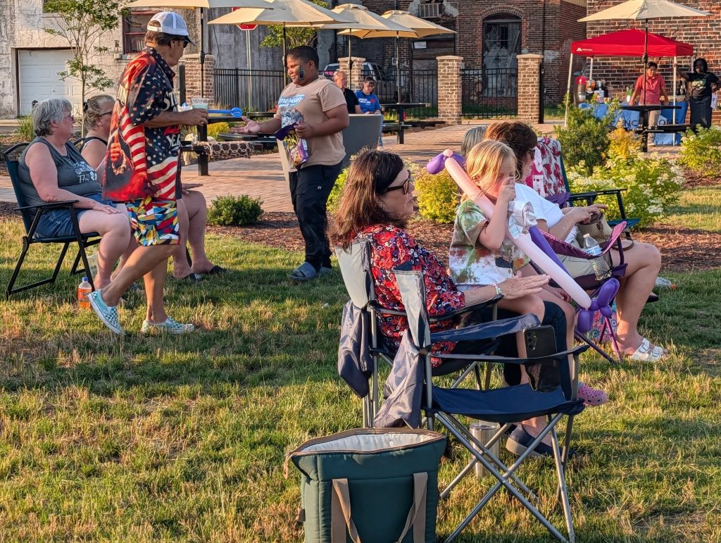 People relaxing on lawn chairs at an outdoor gathering, with others walking nearby. The scene is set in a sunny park with umbrellas and buildings in the background.