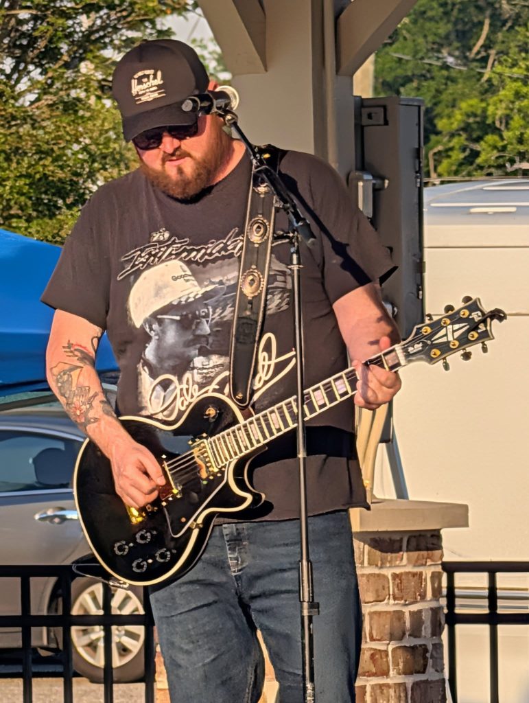A man wearing sunglasses and a hat plays an electric guitar and sings into a microphone during an outdoor performance.