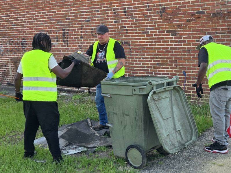 Three people in safety vests clean up debris and place it into a garbage bin near a brick wall outdoors.
