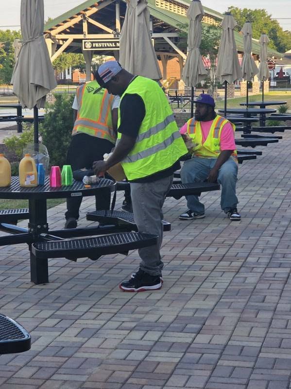 Three workers in safety vests are gathered near outdoor picnic tables with colorful cups and bottles, on a paved patio area with umbrellas.