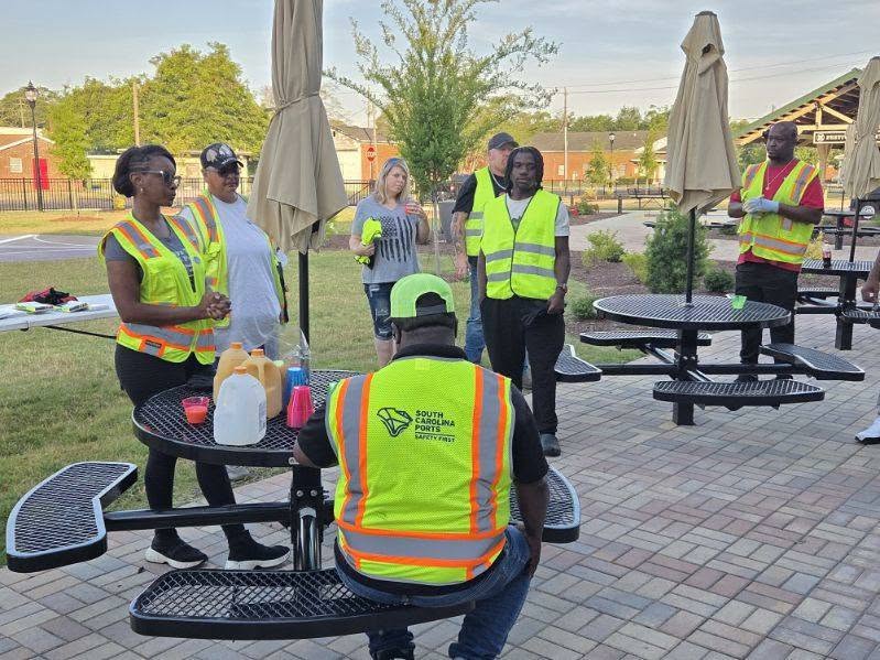 A group of people wearing safety vests gather outdoors around picnic tables, having a meeting or conversation in a paved area with umbrellas.