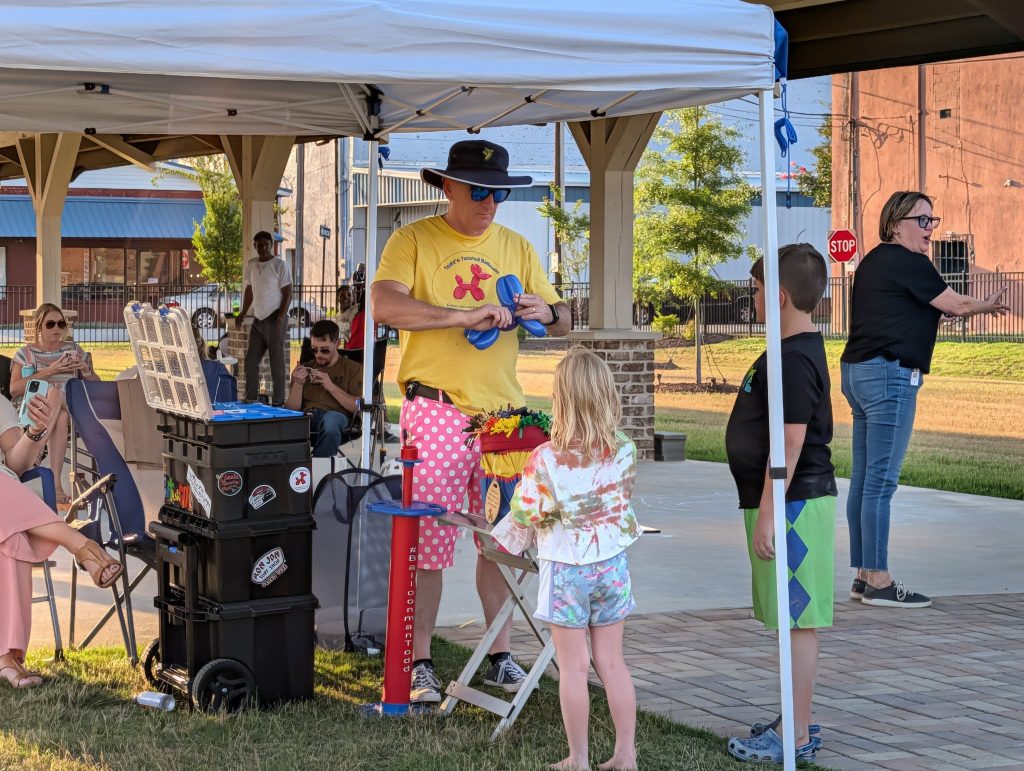 A clown making balloon animals for children under a white tent at an outdoor event, with people and buildings in the background.