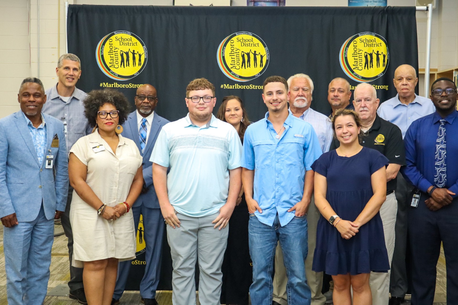 A group of twelve people posing together indoors in front of a black backdrop with yellow logos, some dressed casually and others in business attire.