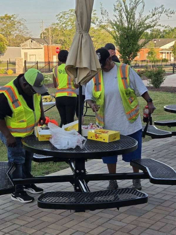 Two workers in safety vests stand at a picnic table with food containers, outdoors in a park setting on a sunny day.