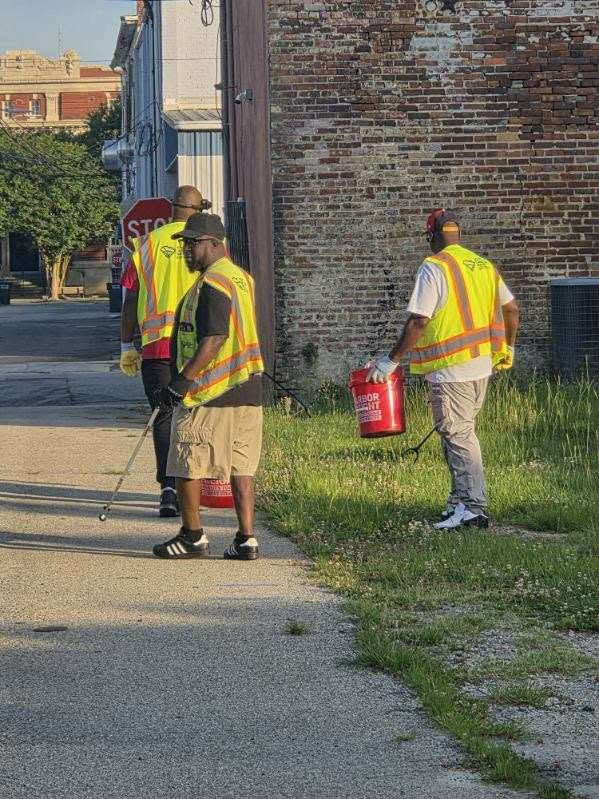 Three workers in yellow safety vests are spraying and carrying red containers while working along a sidewalk near a brick building.