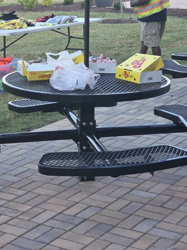 A round outdoor metal picnic table with empty food containers and bags left on top, situated on a paved area with grass in the background.