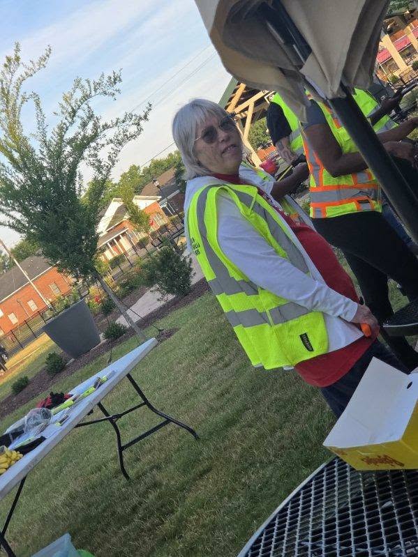 Person wearing a yellow safety vest stands outdoors near tables, with other people in safety vests nearby. Grass, trees, and buildings are visible in the background.