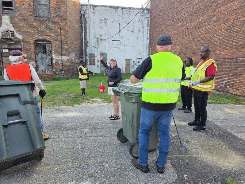 Several people wearing safety vests gather in an alleyway with large trash bins, while one person gestures and others listen near old brick buildings.