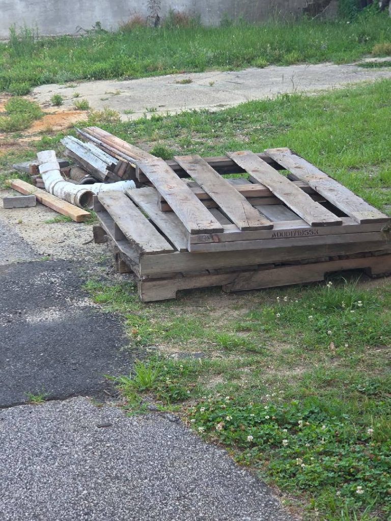 Several wooden pallets stacked on grass near a paved area, with some pipes and wooden planks lying on top and around them.