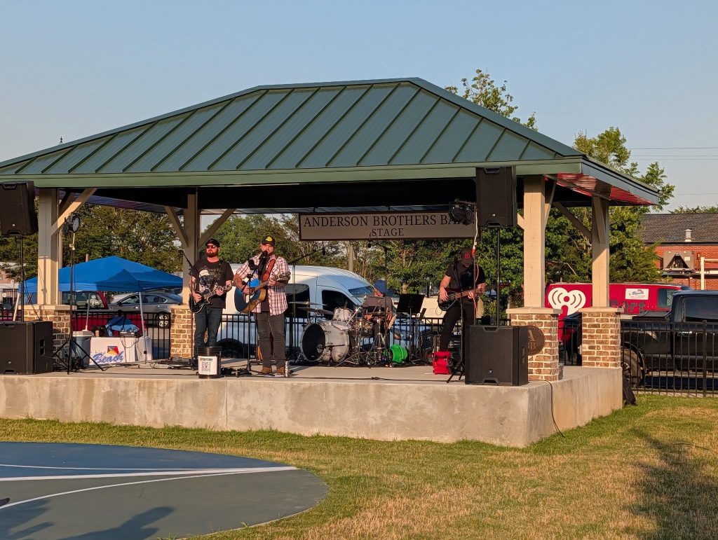 A band performing on an outdoor stage under a pavilion, with musical equipment and audience tents visible in the background on a sunny day.