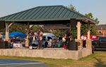 A band performing on an outdoor stage under a pavilion, with musical equipment and audience tents visible in the background on a sunny day.