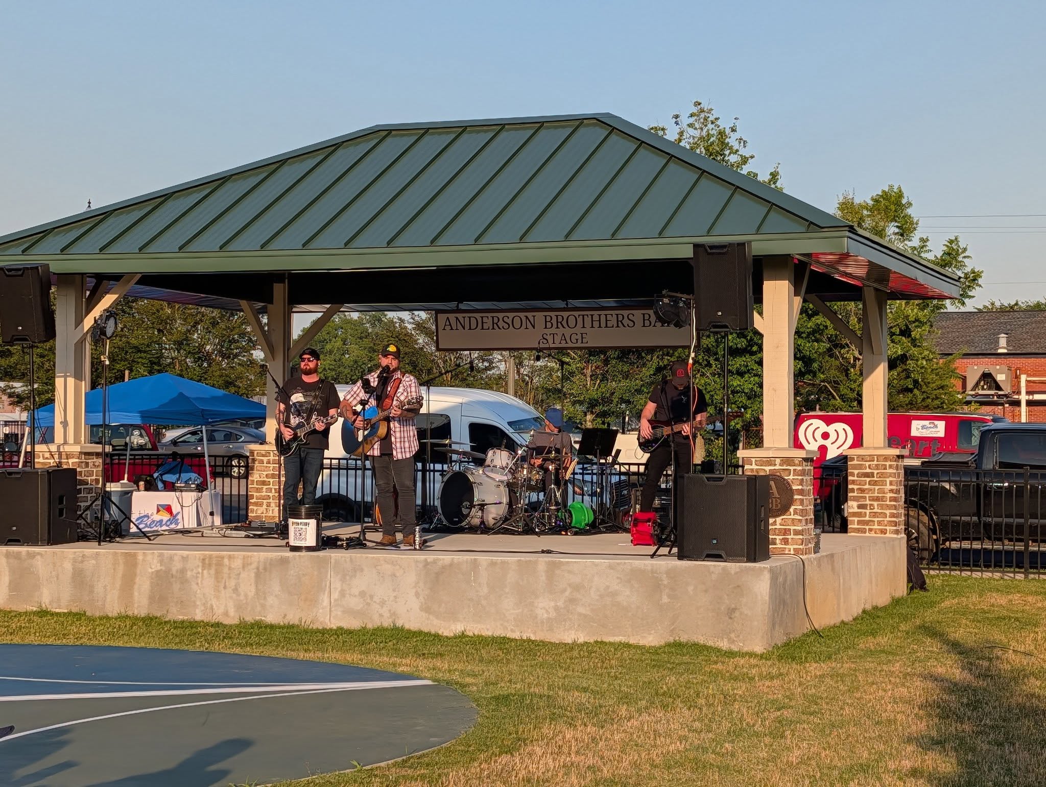A band performing on an outdoor stage under a pavilion, with musical equipment and audience tents visible in the background on a sunny day.