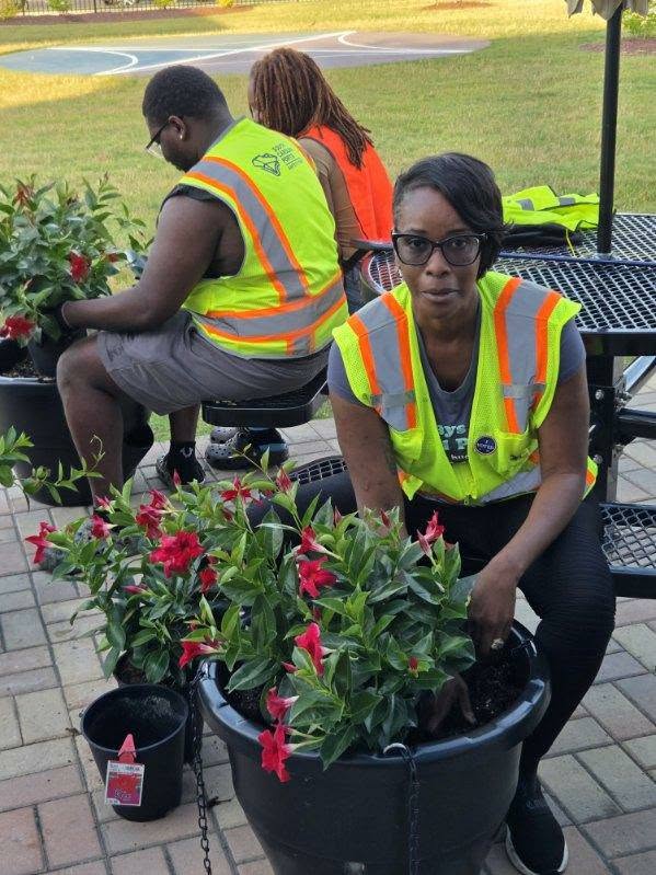 Two people in safety vests sit and work with potted red flowering plants outdoors on a paved area with benches.
