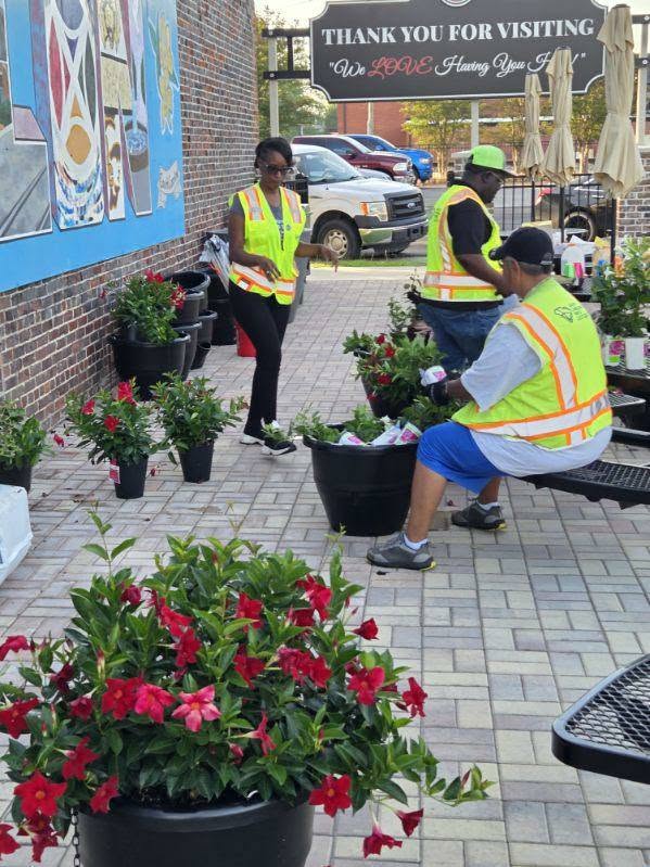 Three people wearing safety vests arrange potted plants outside a building on a brick walkway, with colorful flowers and greenery visible around them.