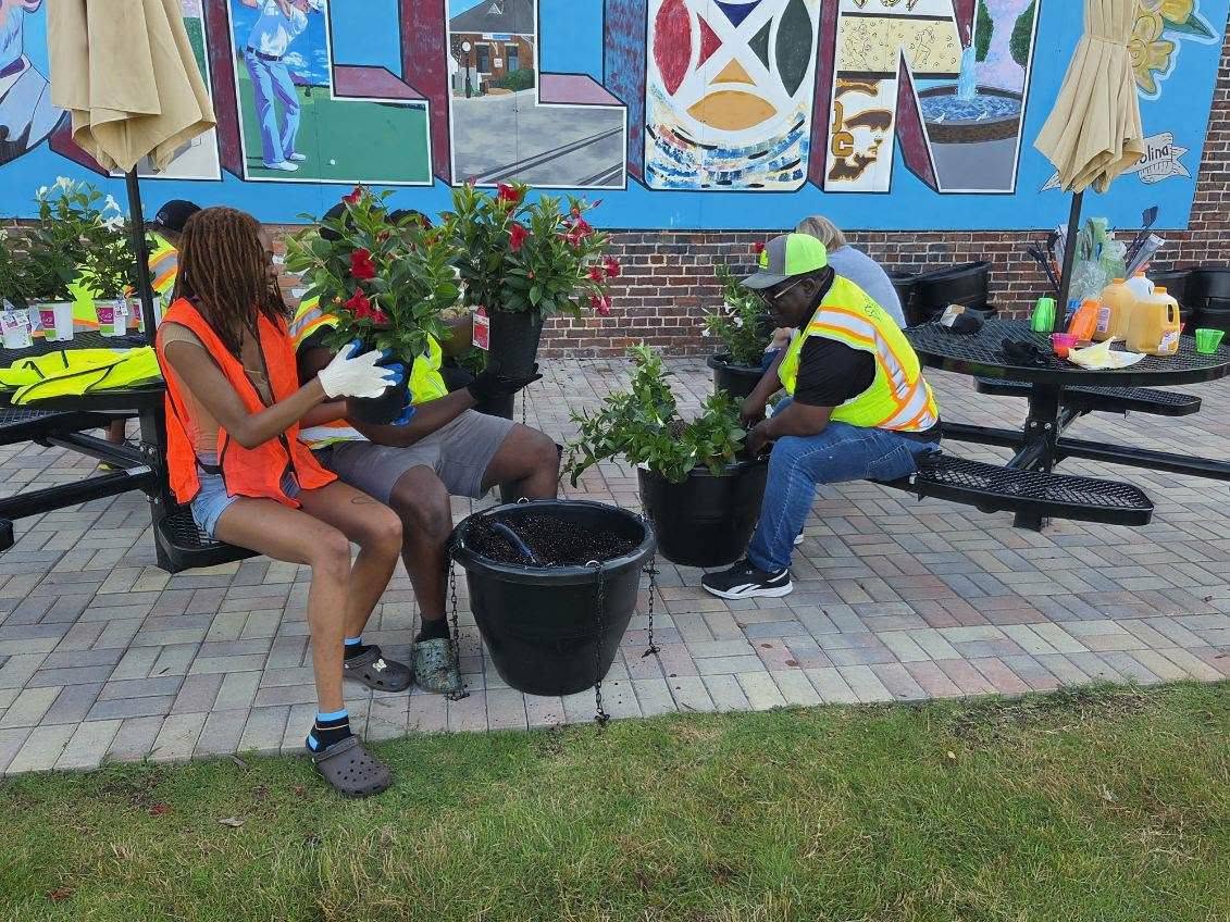 People wearing safety vests sit on benches and plant flowers in pots on a paved area near a colorful mural.