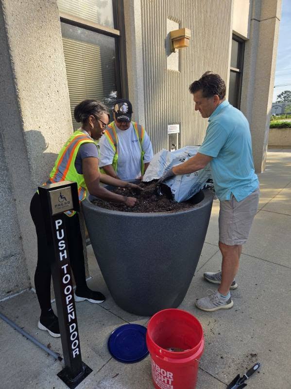 Three people fill a large outdoor planter with soil near a building, using bags of soil and a red bucket nearby.