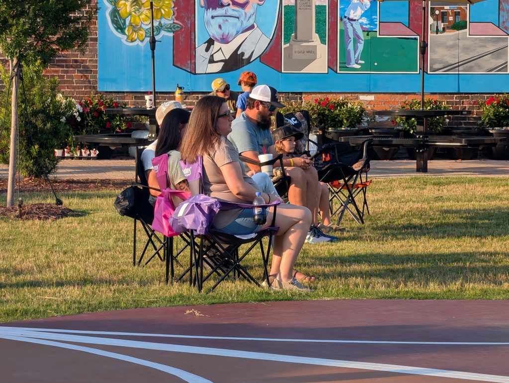 People sitting on portable chairs in a grassy park near a basketball court, with a colorful mural visible on a wall in the background.