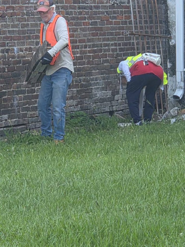 Two workers in safety vests are working near a brick wall, one standing with equipment, the other bending down near the foundation, with grass in the foreground.