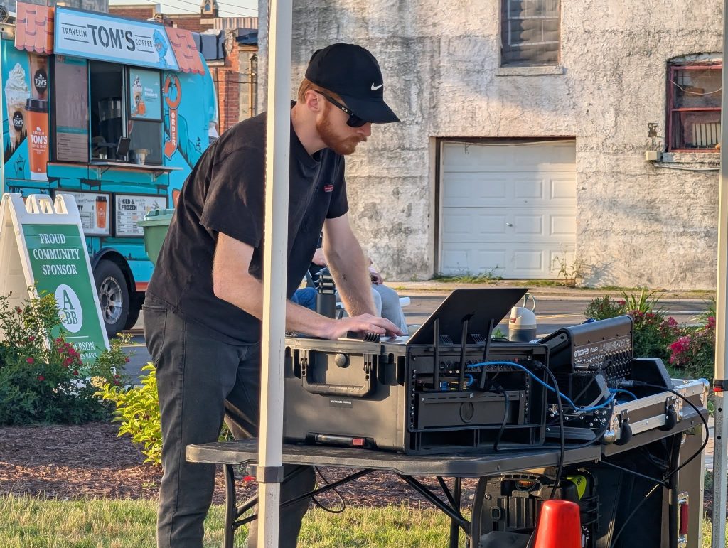 A person wearing black operates DJ equipment outdoors near a food truck, standing at a table with audio gear and a laptop.