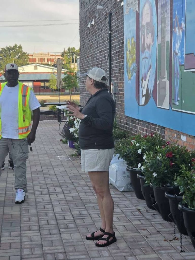 A person in a safety vest stands outside talking to another person near a mural and potted plants lined along a brick wall.