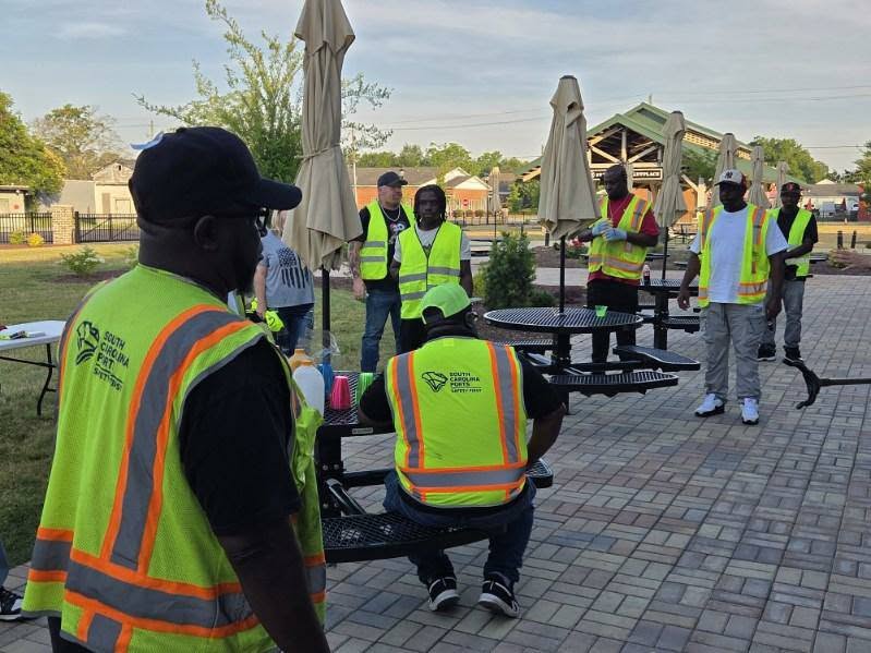A group of people wearing yellow safety vests gather outdoors near picnic tables and umbrellas on a paved area, possibly preparing for work or a community event.