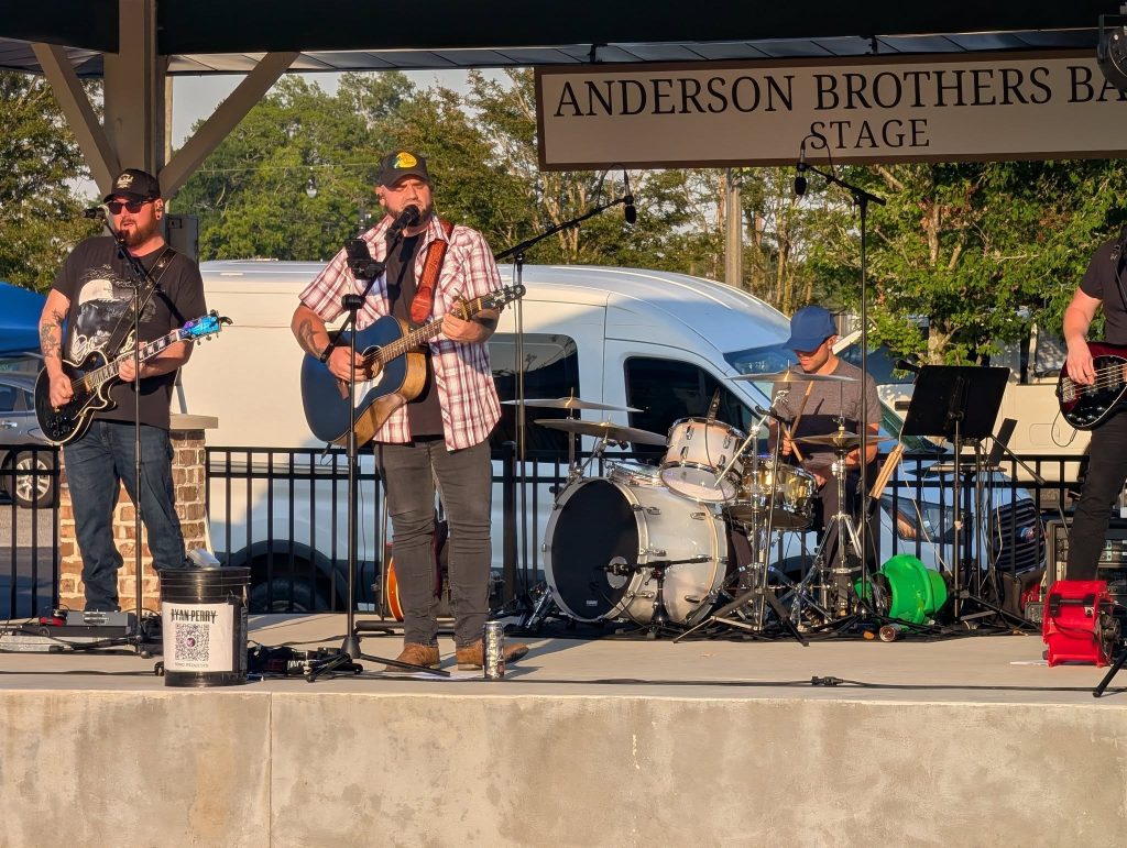A band performs on an outdoor stage under a banner reading "Anderson Brother Stage," with guitars and drums visible, and a van parked in the background.