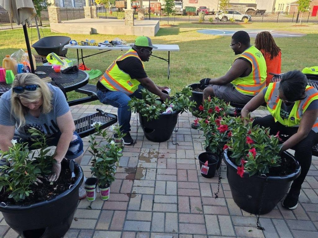 Four people wearing safety vests are sitting outside, planting flowers in large pots on a paved surface. Gardening tools and supplies are on nearby tables.