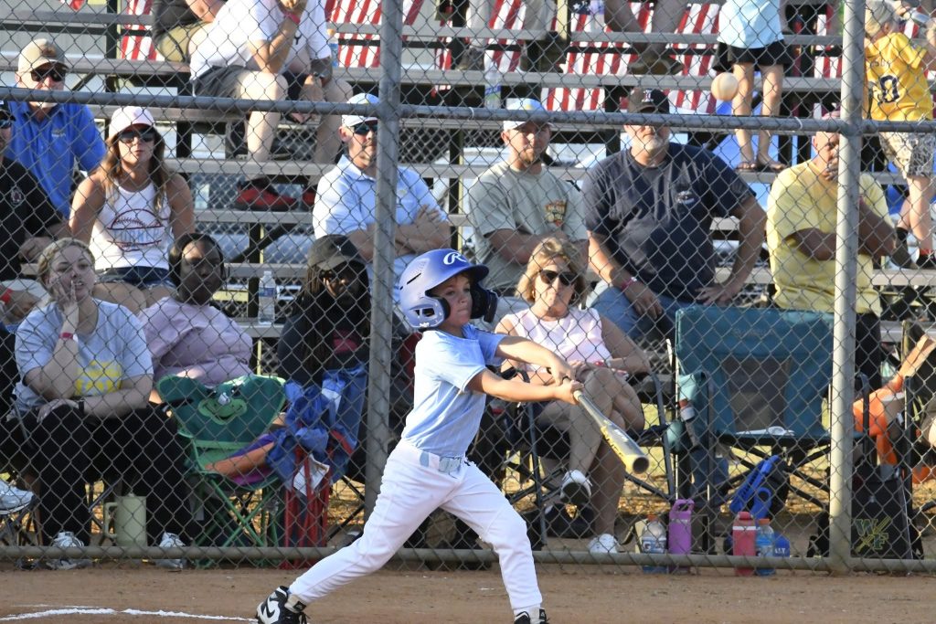 A young baseball player swings a bat during a game, with spectators watching from bleachers behind a chain-link fence.