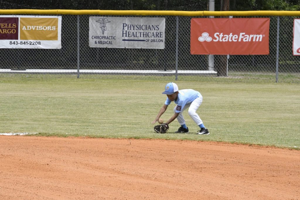 A young baseball player in a light blue uniform fields a ground ball on a grassy field near the infield dirt.