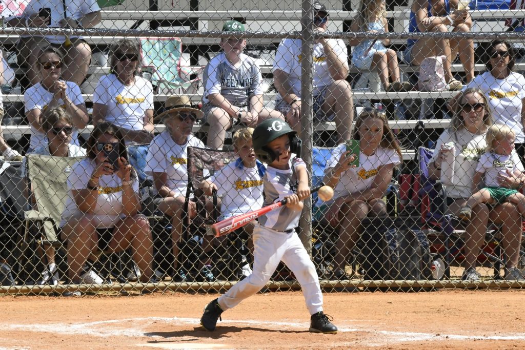 A young child wearing a helmet swings a bat at a softball game, with spectators watching from bleachers behind a chain-link fence.
