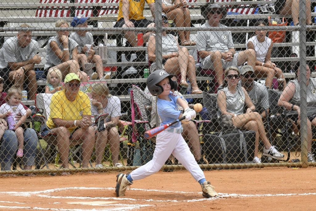 A young baseball player swings a bat during a game, with spectators watching from bleachers behind a chain-link fence.