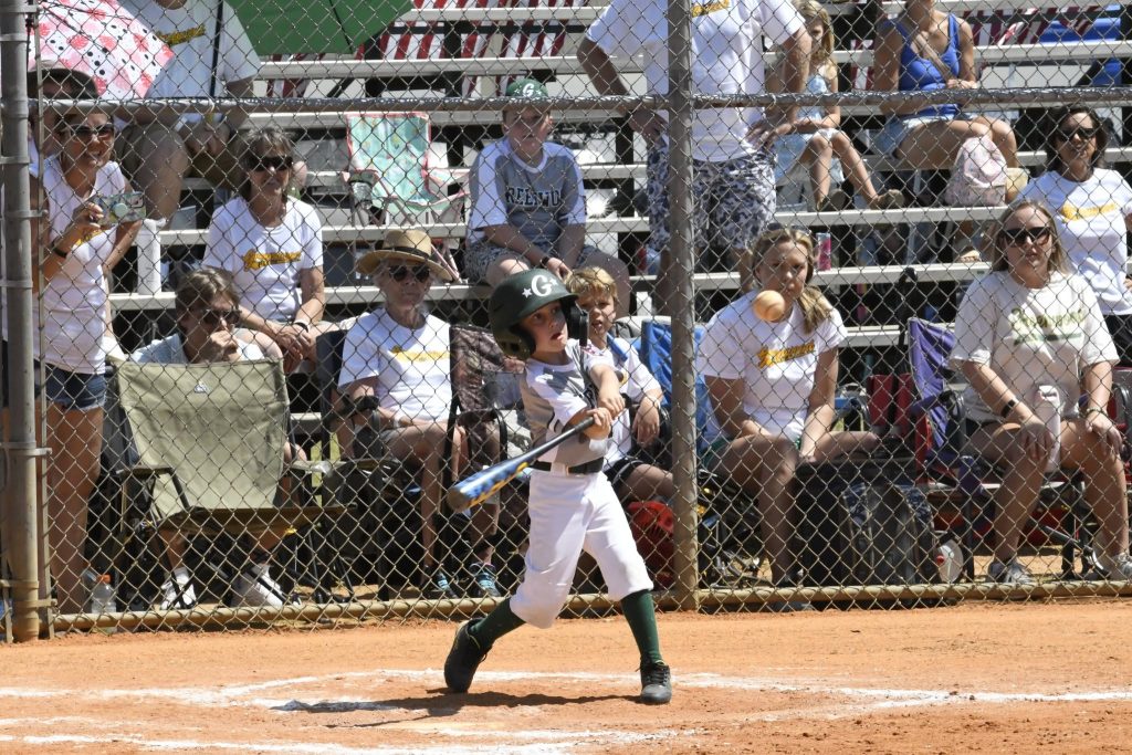 A young baseball player swings a bat during a game, with spectators watching from bleachers behind a chain-link fence.