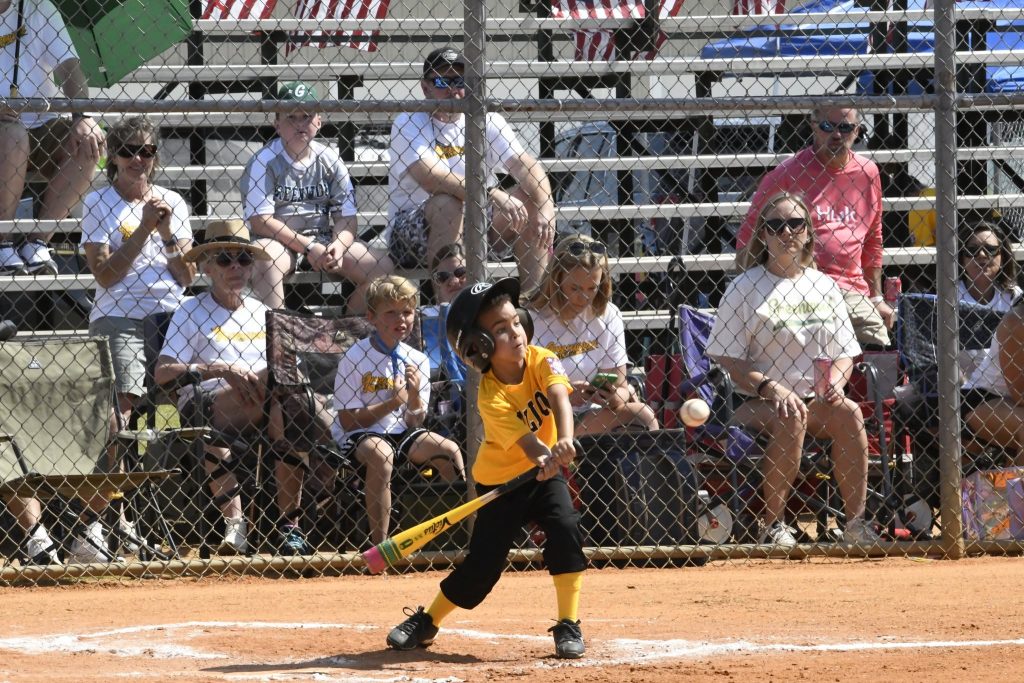 A young softball player in a yellow jersey swings a bat, with spectators watching from bleachers behind a chain-link fence on a sunny day.