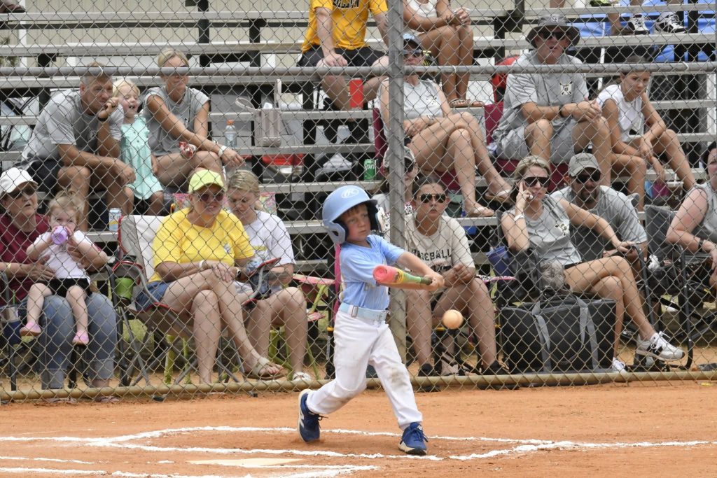 Young child in blue helmet swinging a baseball bat during a game, with spectators watching from bleachers behind a chain-link fence.