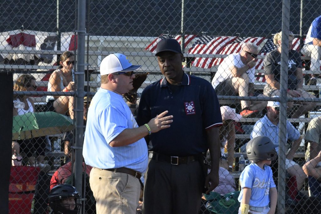 Two men, one in a light shirt and cap, the other in a dark uniform, talking on a baseball field with spectators in the background.