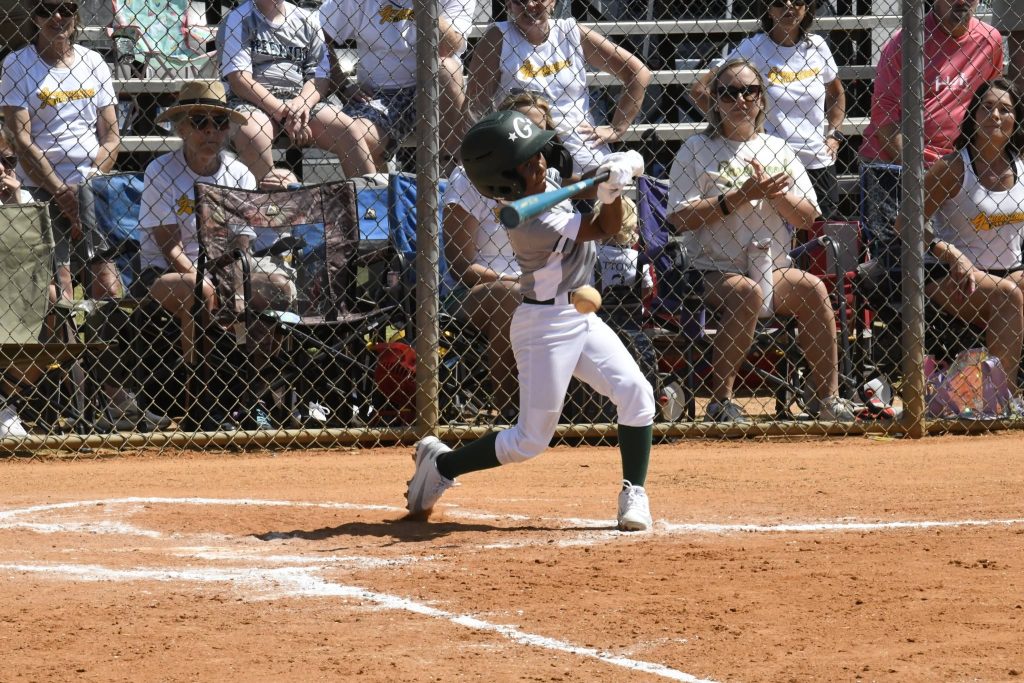 A young baseball player in white uniform swinging a bat at home plate, with spectators watching from bleachers in the background.
