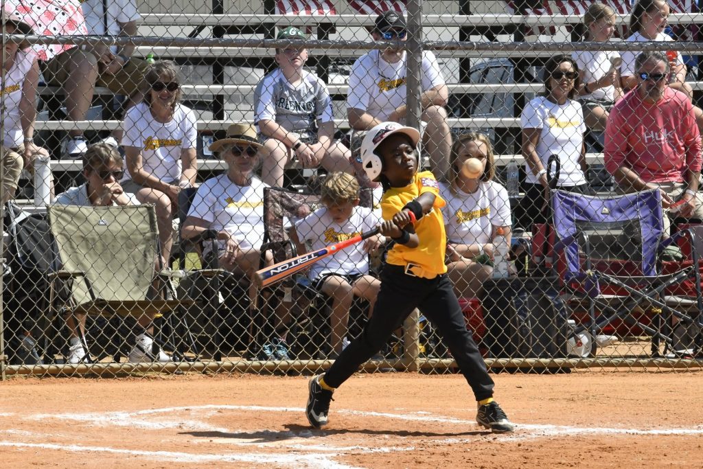 A young baseball player in a yellow jersey swings a bat during a game, with spectators watching from bleachers behind a chain-link fence.
