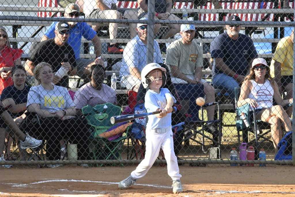 A young child wearing a helmet swings a bat at a baseball game, with spectators watching from the bleachers behind a chain-link fence.