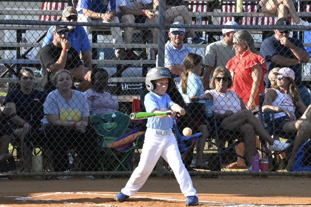 A young child in a helmet swings a bat at a baseball during a game, with spectators watching from the bleachers in the background.