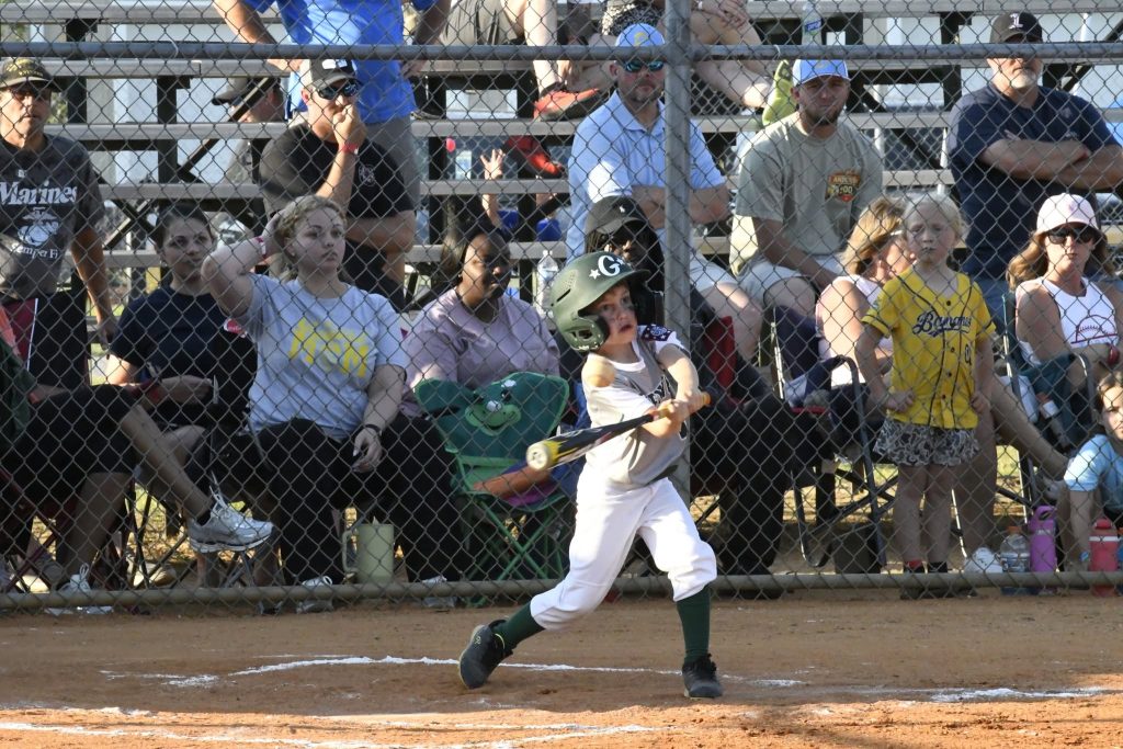 A young child in a helmet swings a baseball bat during a game while spectators watch from bleachers behind a fence.
