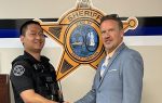 Police officer and man in a suit shake hands in front of a sheriff's office emblem on the wall.