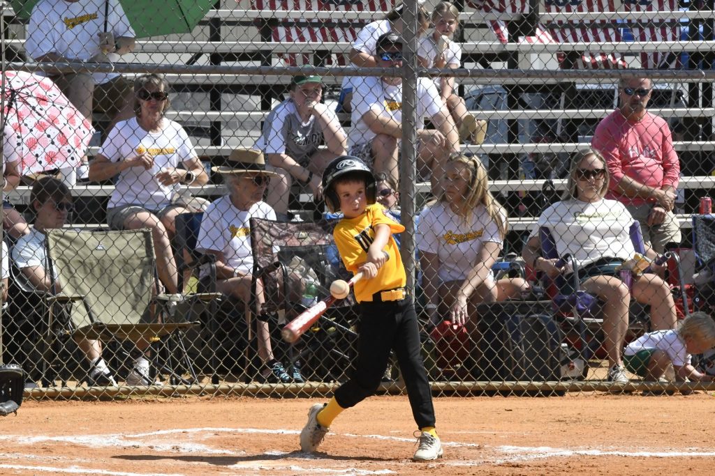A young child in a yellow jersey swings a baseball bat during a game, with spectators watching from bleachers in the background.