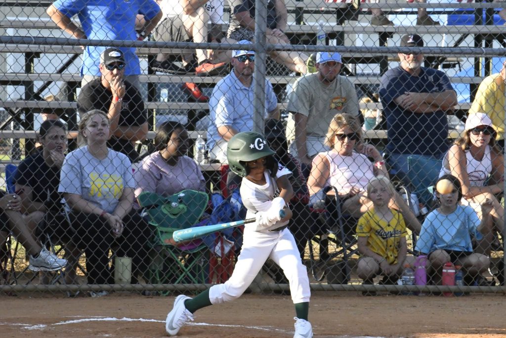 A young baseball player swings a bat during a game, with a crowd of spectators watching from bleachers behind a chain-link fence.