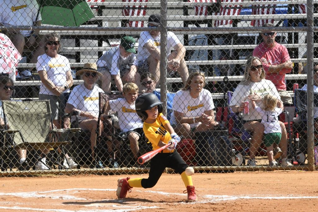 A young child swings a bat during a baseball game, with spectators watching from bleachers in the background.