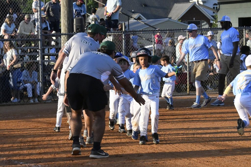 Young children in baseball uniforms high-five coaches on a dirt field, with spectators watching behind a fence.