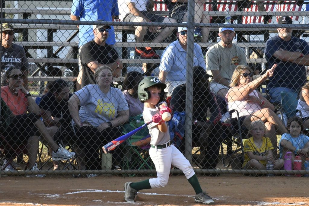 A young child in baseball gear swings a bat during a game, with spectators watching from the bleachers behind a chain-link fence.