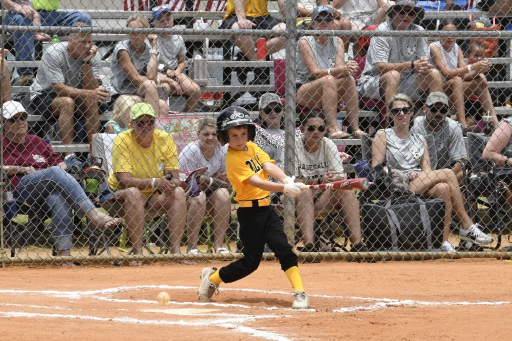 A young baseball player in a yellow jersey swings at a pitch during a game, with spectators watching from the bleachers behind a chain-link fence.